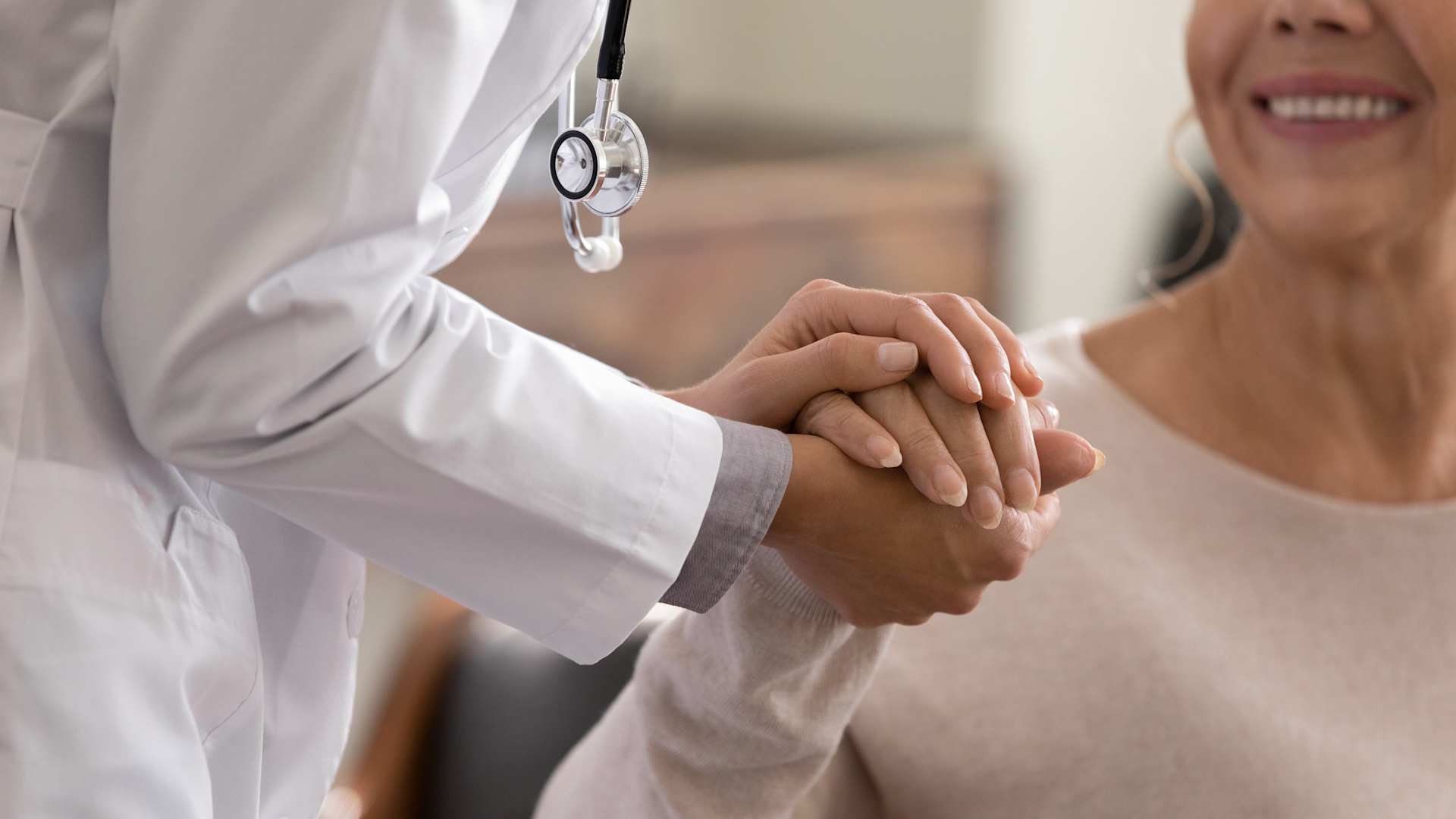 Doctor in white coat with stethoscope holding a smiling female patient's hands in a compassionate gesture